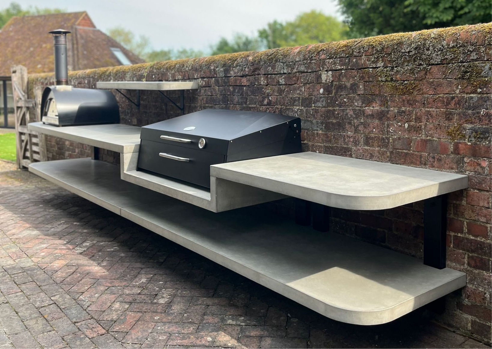 Pink worktop Custom multi-level concrete outdoor kitchen featuring a built-in pizza oven and integrated BBQ grill station against a rustic brick wall.