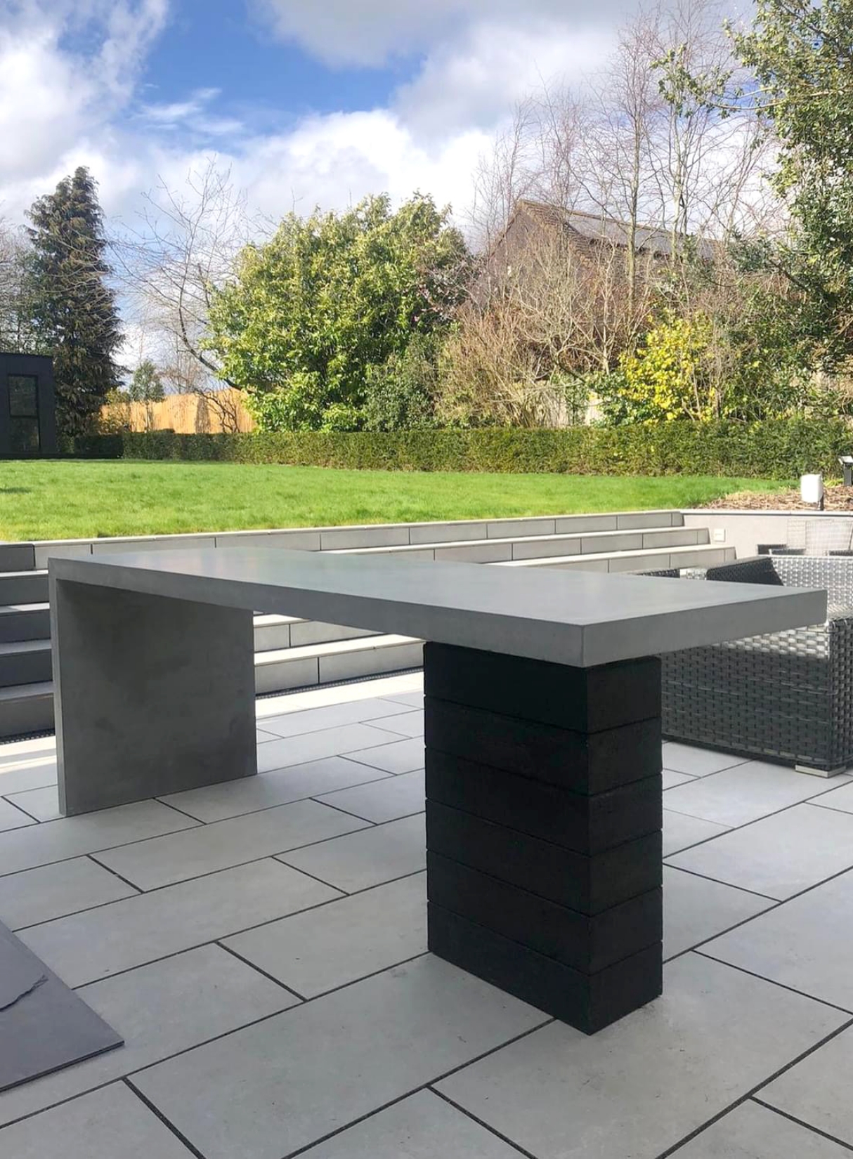 Rectangle 1208 Architectural grey concrete outdoor bar table with a thick waterfall edge, supported by a stacked black block pedestal on a modern grey paved patio.