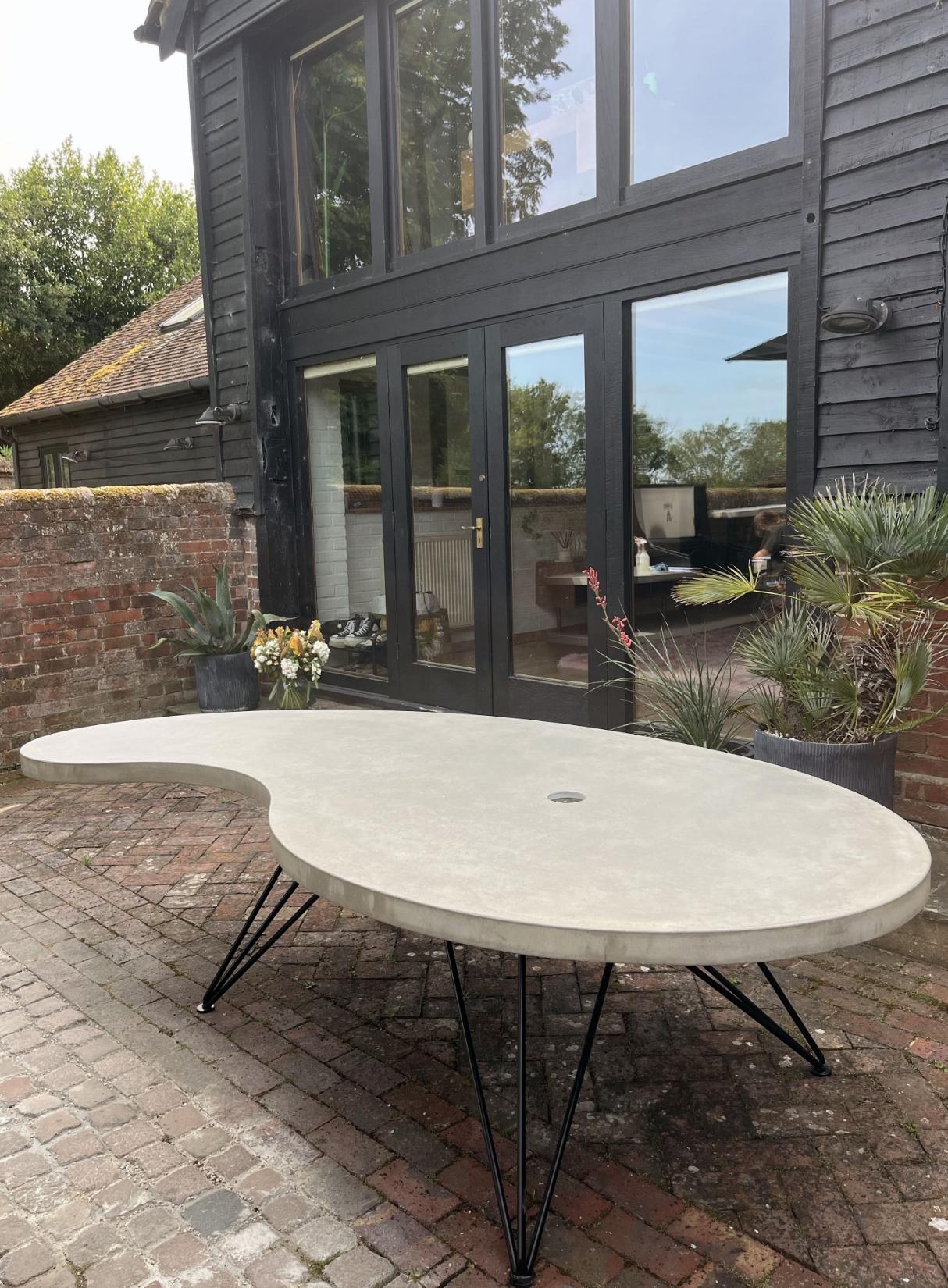 Rectangle 1236 Large bespoke kidney-shaped concrete outdoor dining table with black hairpin legs, featuring a central parasol hole on a rustic brick patio.