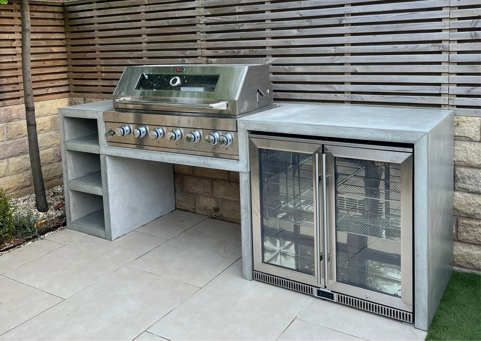 Screenshot 2023-12-19 at 09.16.51 Custom grey concrete outdoor kitchen suite featuring a high-performance integrated gas BBQ grill, built-in shelving, and a double-door outdoor drinks fridge under a seamless concrete worktop.
